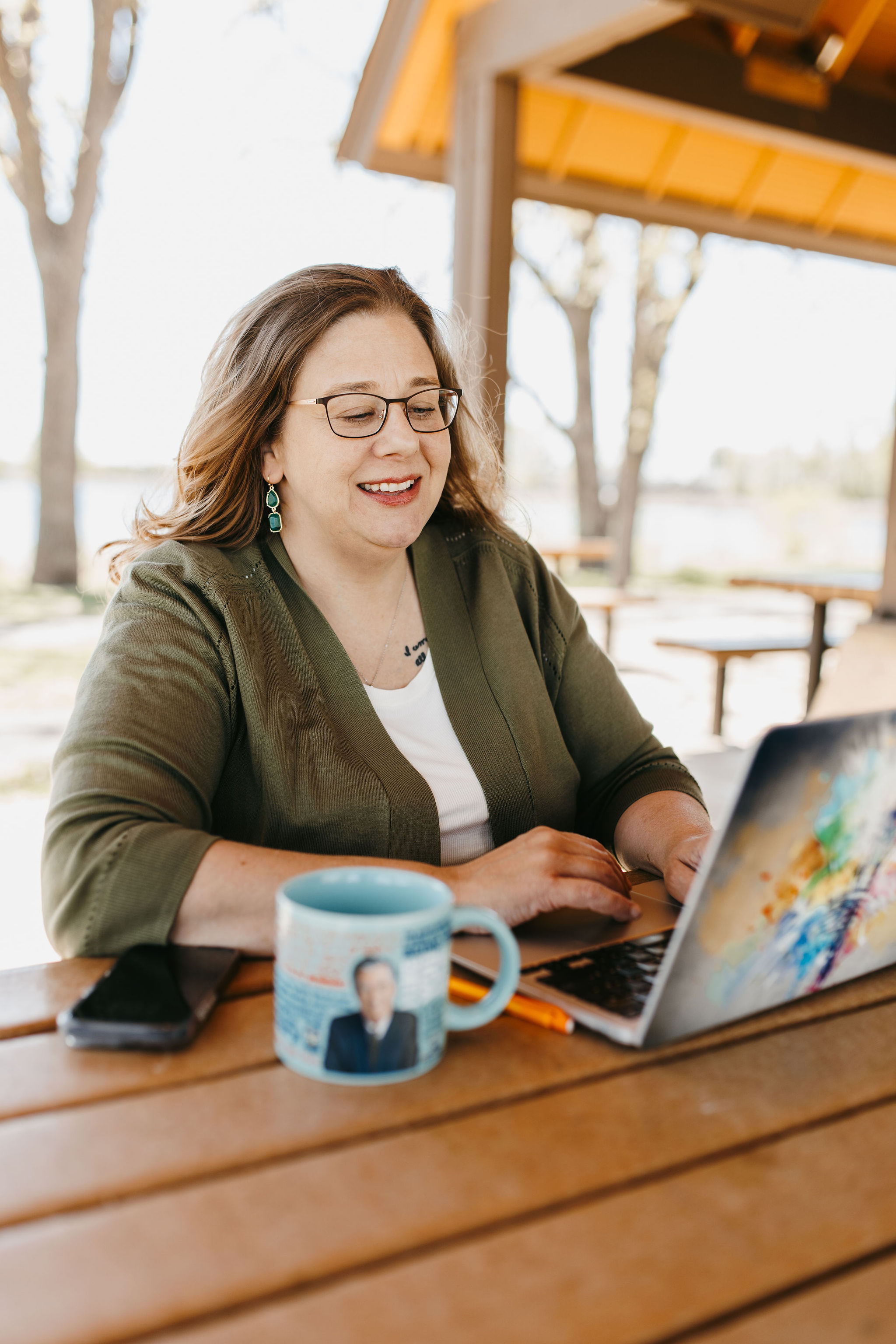 woman with dark hair in green sweater working on laptop with coffee mug at picnic table outside