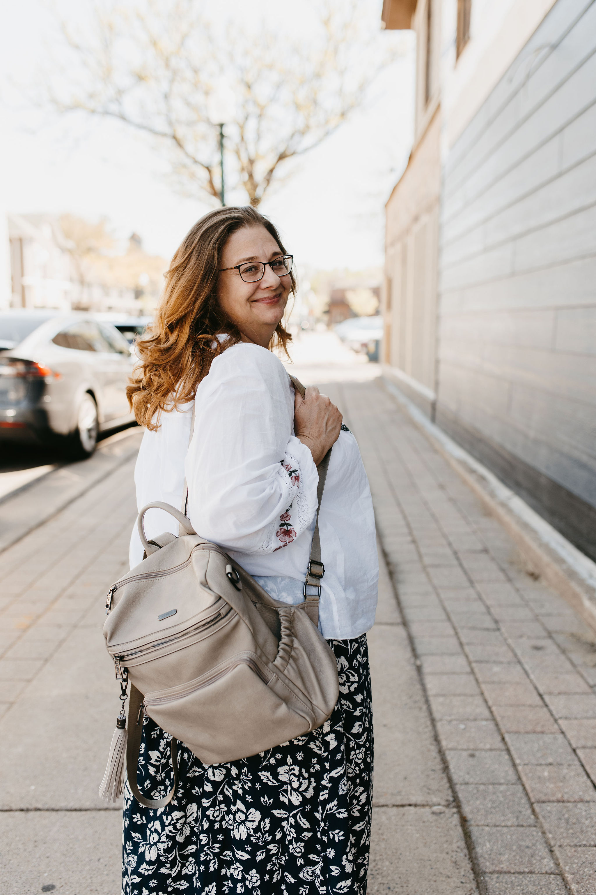 woman with redish dark hair outside in white blouse and black floral skirt and grey backpack