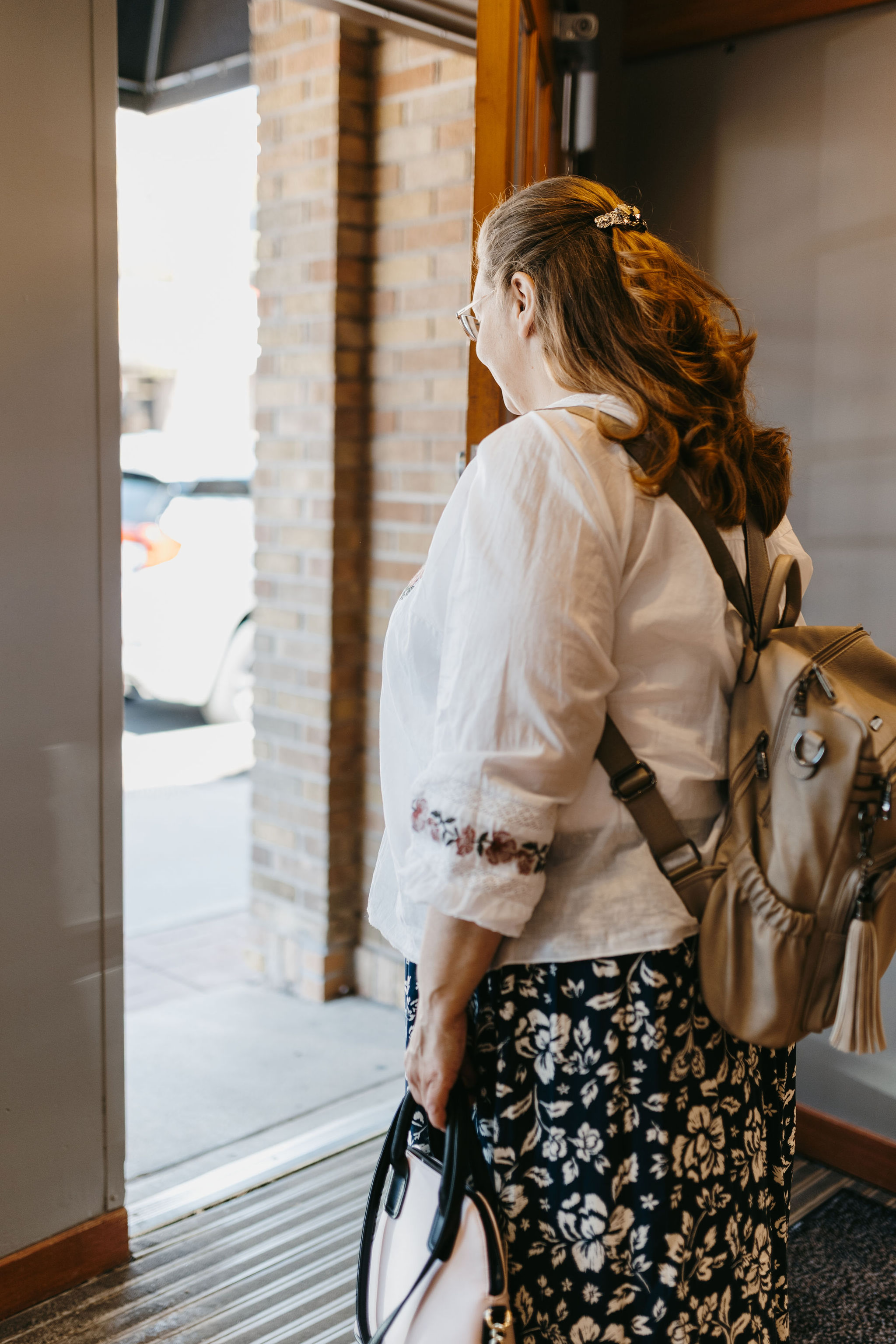 woman with dark reddish hair walking brick doorway in white blouse and black floral skirt with backpack